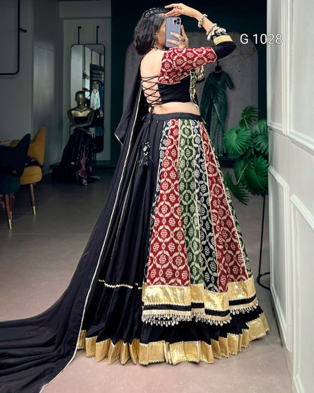 A woman takes a mirror selfie in a boutique wearing a traditional Indian lehenga choli outfit. The outfit features a black base with intricate red, green, and gold patterns on the skirt, a red and gold patterned blouse, and a long black dupatta with gold borders.