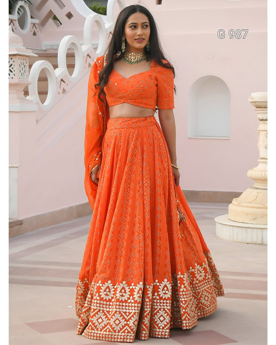 A woman modeling a bright orange embroidered Indian ethnic outfit with a flared skirt, short sleeves, and a matching dupatta, standing in front of a pale pink architectural background.