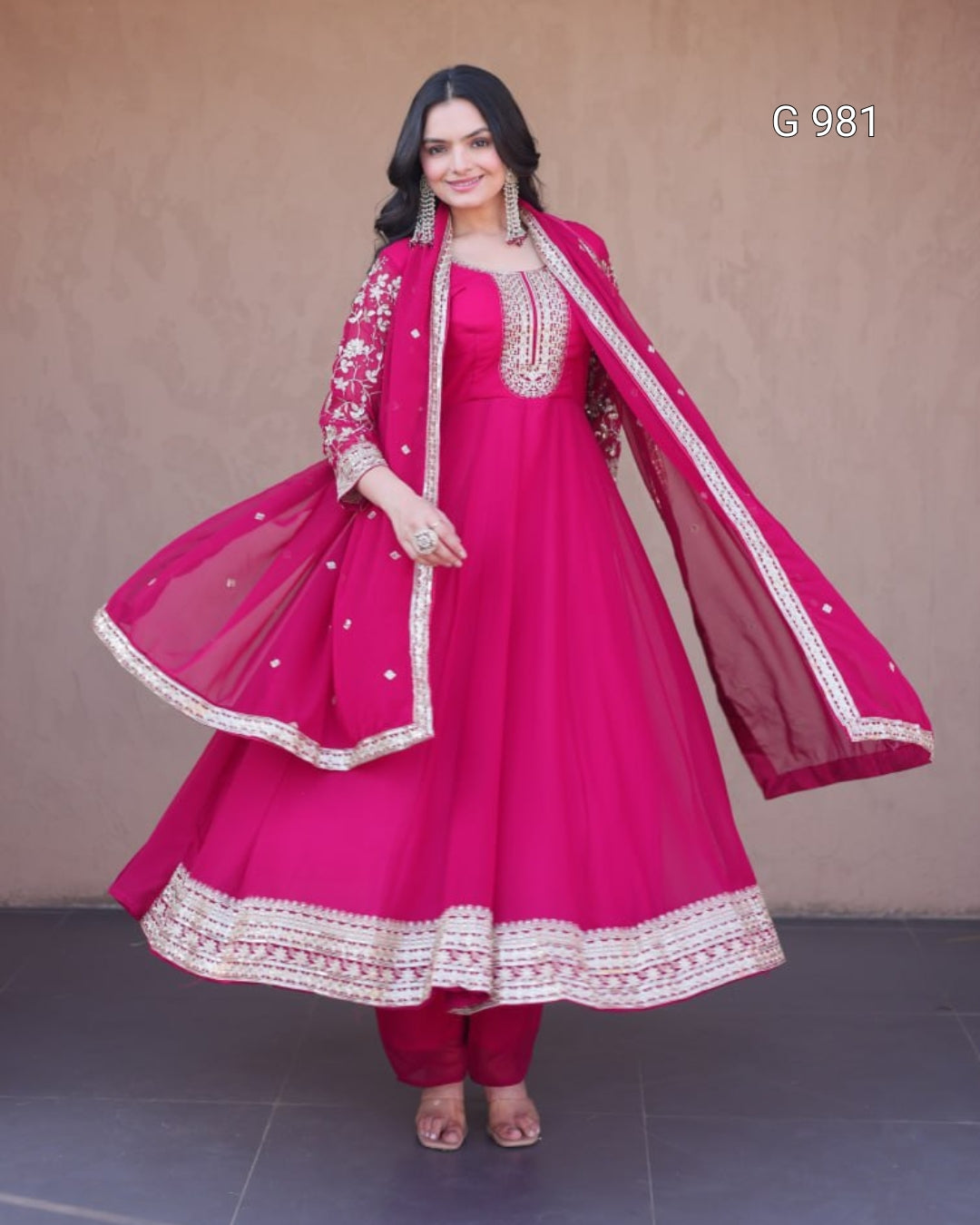 A woman modeling a bright pink Indian ethnic outfit, featuring a heavily embroidered anarkali kurta, matching pants, and a sheer dupatta draped over her shoulders, set against a plain brown wall.