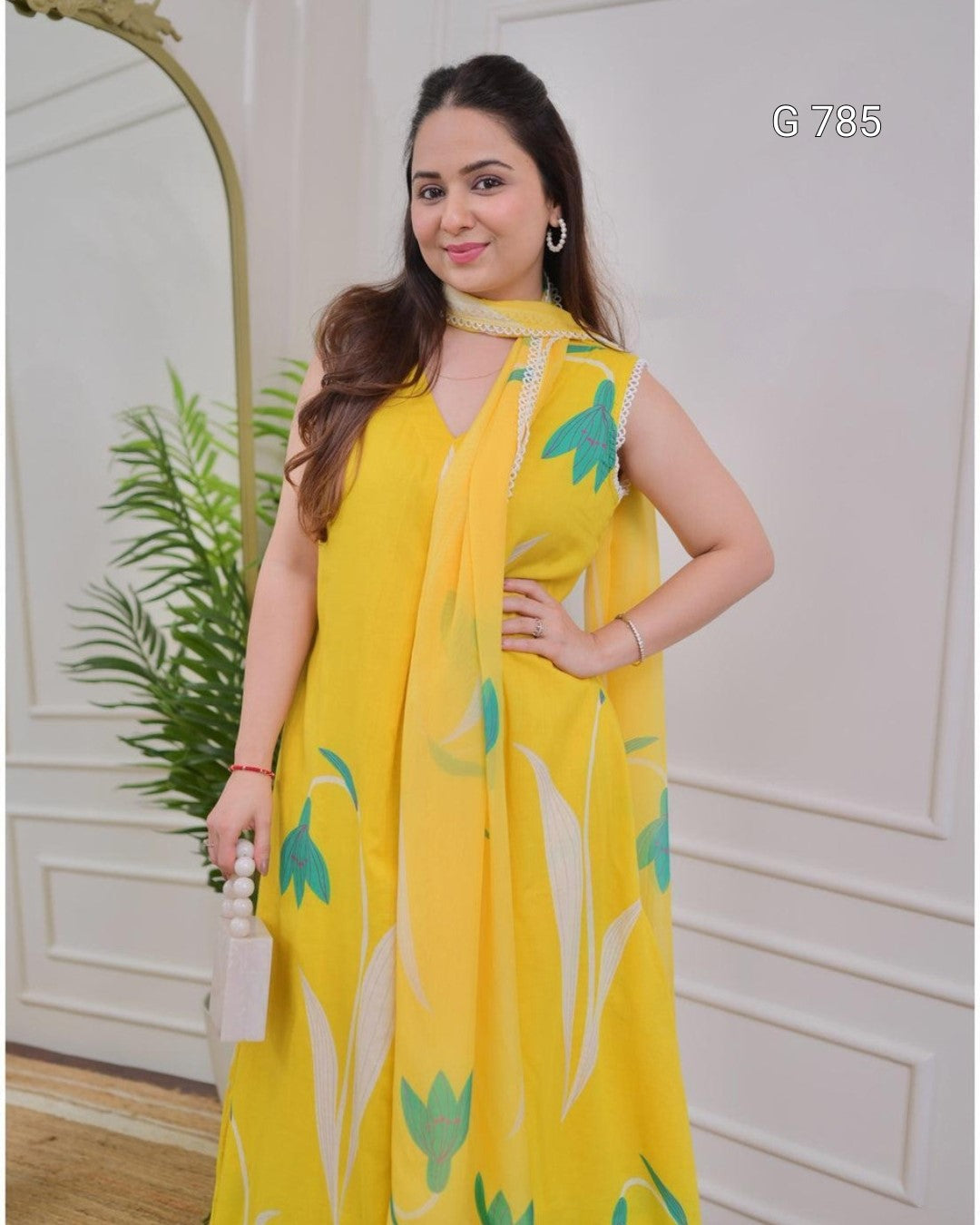 Woman wearing a yellow floral dress standing indoors with a decorative mirror and plant in the background.