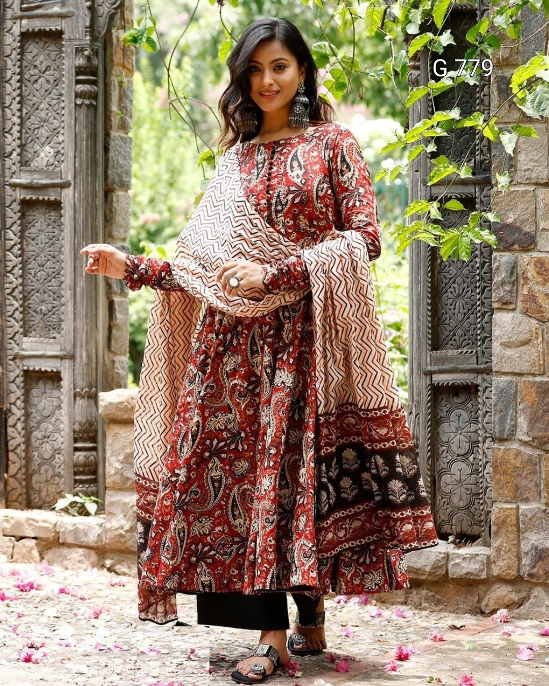 Woman in a patterned dress standing in a decorative outdoor setting with stone architecture and greenery.