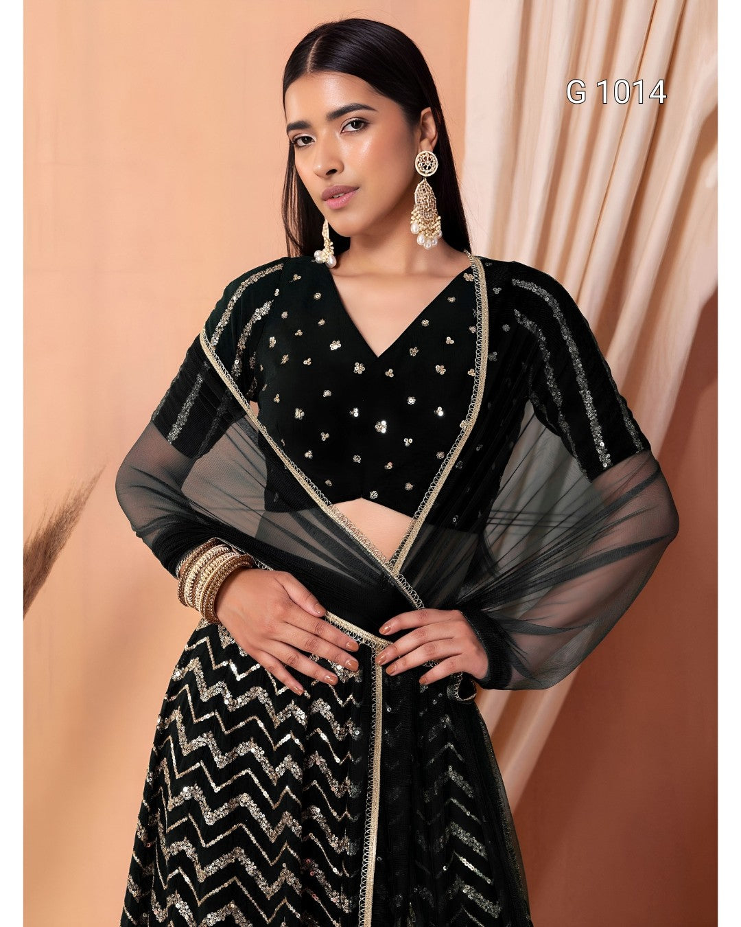 A woman modeling a black traditional outfit with silver embellishments, featuring a patterned skirt, sheer sleeves, and a matching dupatta, posed against a peach-colored backdrop.