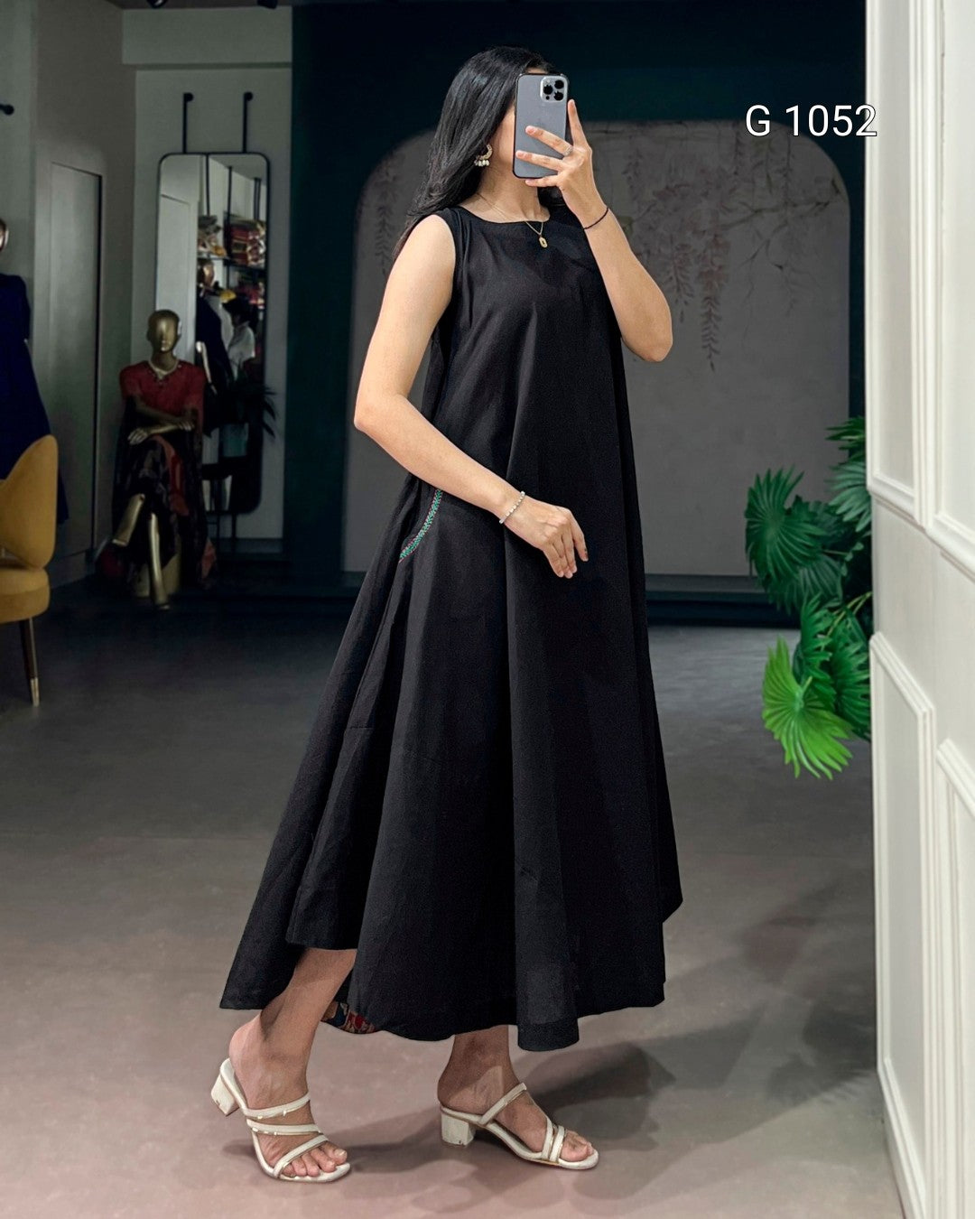 A woman takes a mirror selfie in a boutique wearing a sleeveless black midi dress with a flared skirt and white strappy heeled sandals. The boutique interior has a gray floor, a mannequin in the background, and a green plant on the right.
