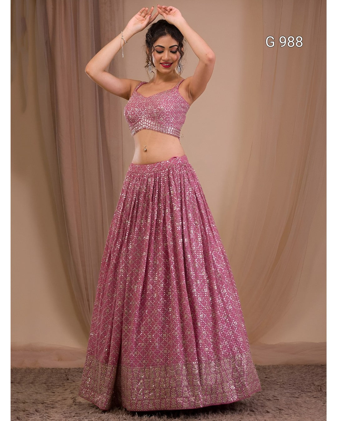 A woman modeling a pink embellished lehenga choli set against a beige draped backdrop. The set features a pink sequined crop top and a matching flared skirt with a decorative border.