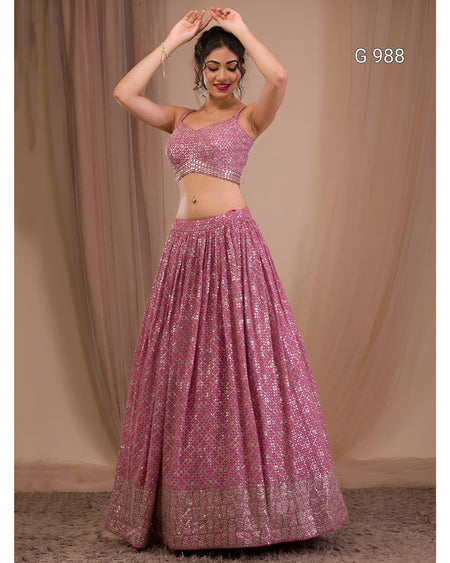 A woman modeling a pink embellished lehenga choli set against a beige draped backdrop. The set features a pink sequined crop top and a matching flared skirt with a decorative border.