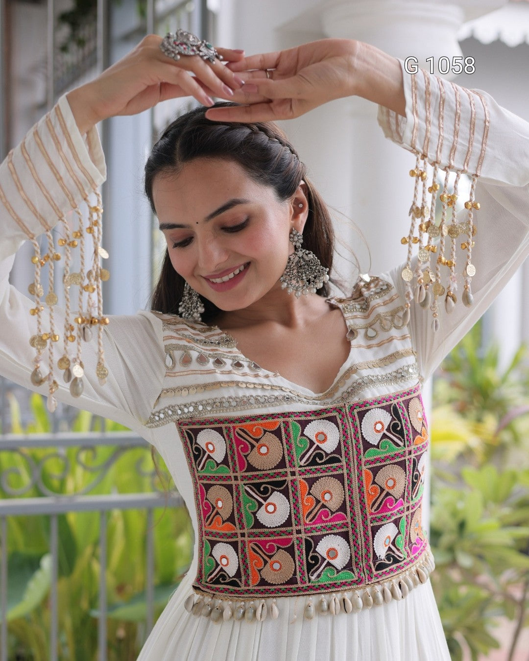 A woman wearing a white embroidered traditional dress with colorful geometric patterns on the bodice and gold coin tassels on the sleeves, posing with her hands near her head in an outdoor setting.