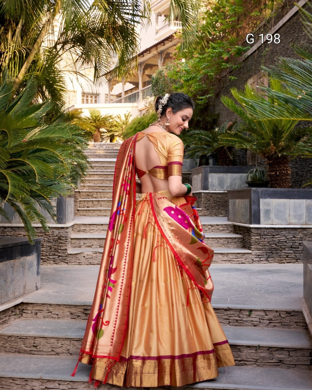 Woman in a traditional saree standing outdoors with greenery and steps in the background