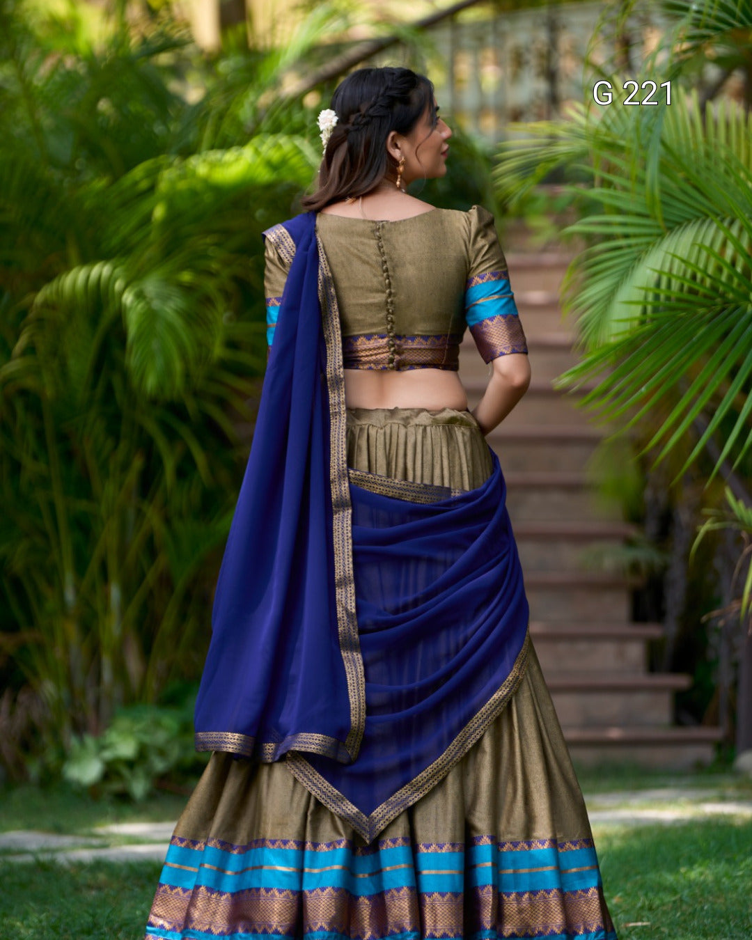Woman in traditional green and blue saree standing outdoors with greenery in the background