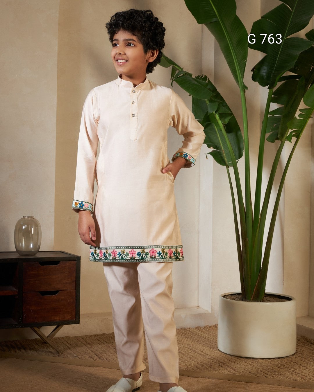 Young boy in a traditional outfit standing indoors with a plant in the background