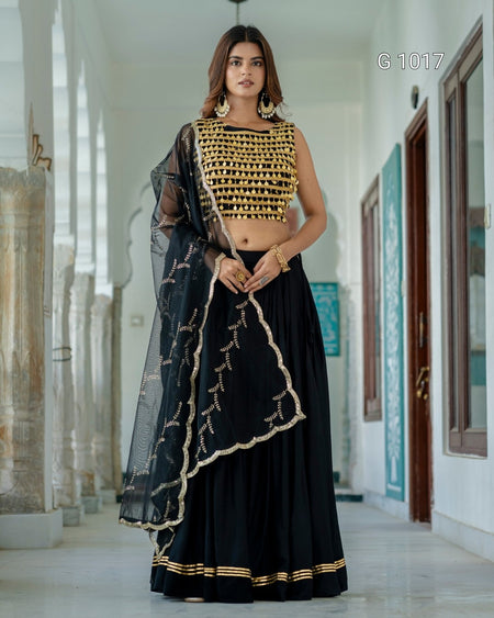 A woman models a traditional Indian outfit consisting of a black skirt with gold embroidery, a gold embroidered sleeveless crop top, and a black sheer dupatta with gold borders, set against a white corridor background.