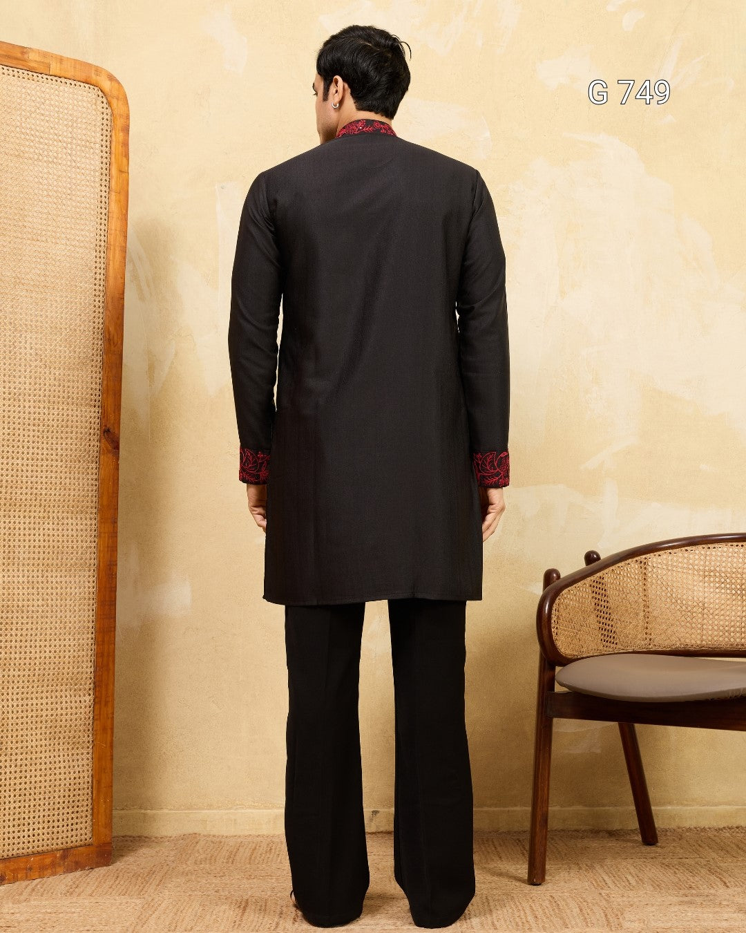 Man wearing a black traditional outfit with red accents in a room with a chair and woven screen.