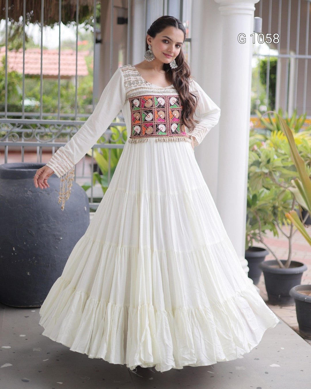 A woman models a long white Anarkali gown with a heavily embroidered multicolor bodice and a tiered skirt, standing on a porch with potted plants in the background.