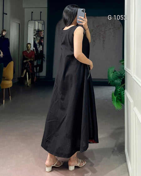 A woman takes a mirror selfie in a boutique wearing a sleeveless black maxi dress with a gathered waist and tie detail, paired with beige block-heel mules. The boutique interior features a gray floor, a dark textured wall, and mannequins in the background.
