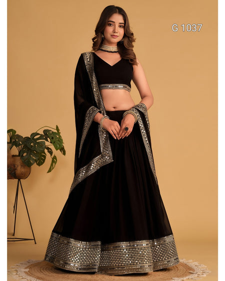 A woman models a black traditional Indian lehenga choli set with silver mirror work borders, standing against a solid beige background.