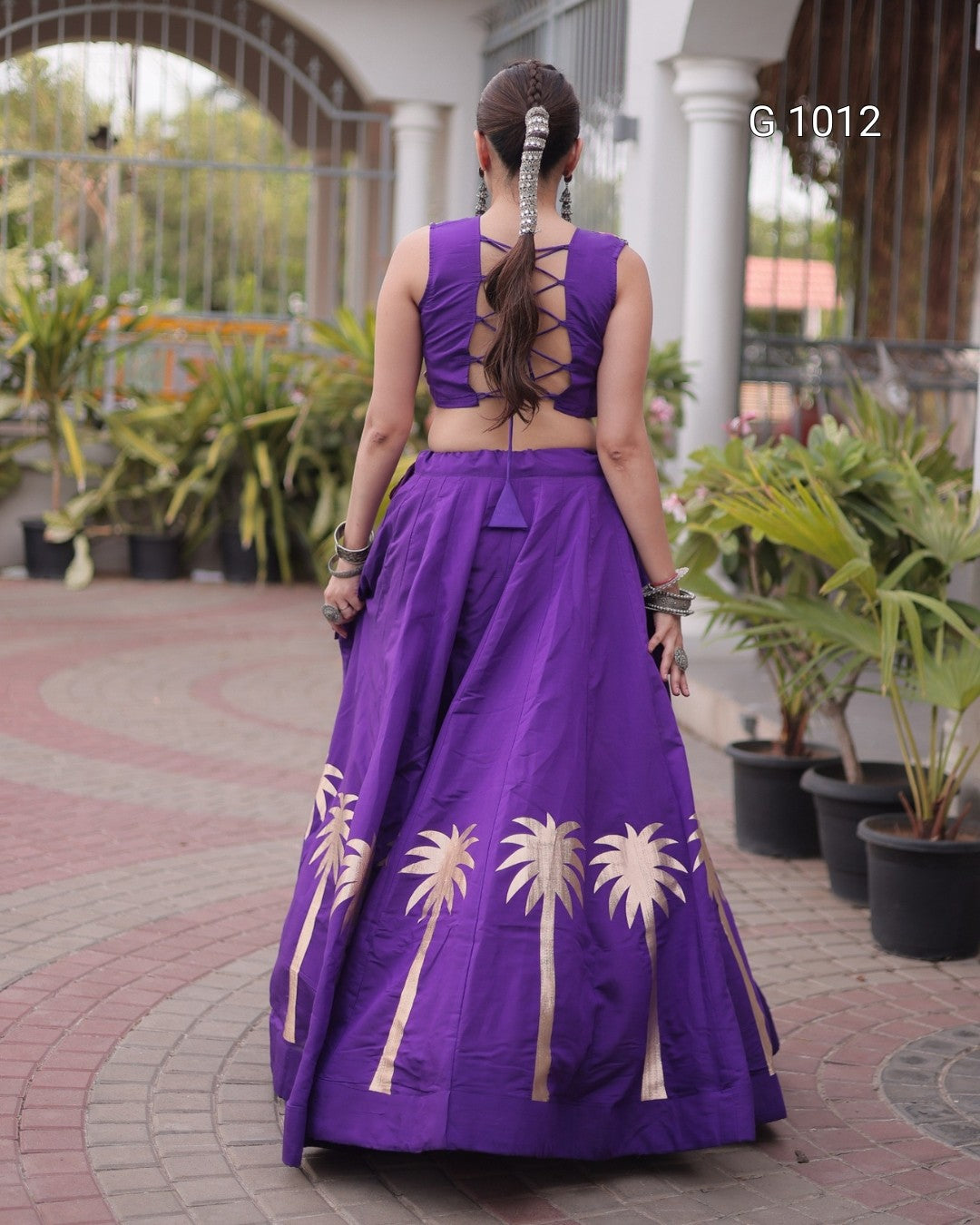 A woman viewed from behind wearing a purple sleeveless lehenga choli set with cream palm tree embroidery on the skirt, standing on a paved walkway with potted plants in the background.