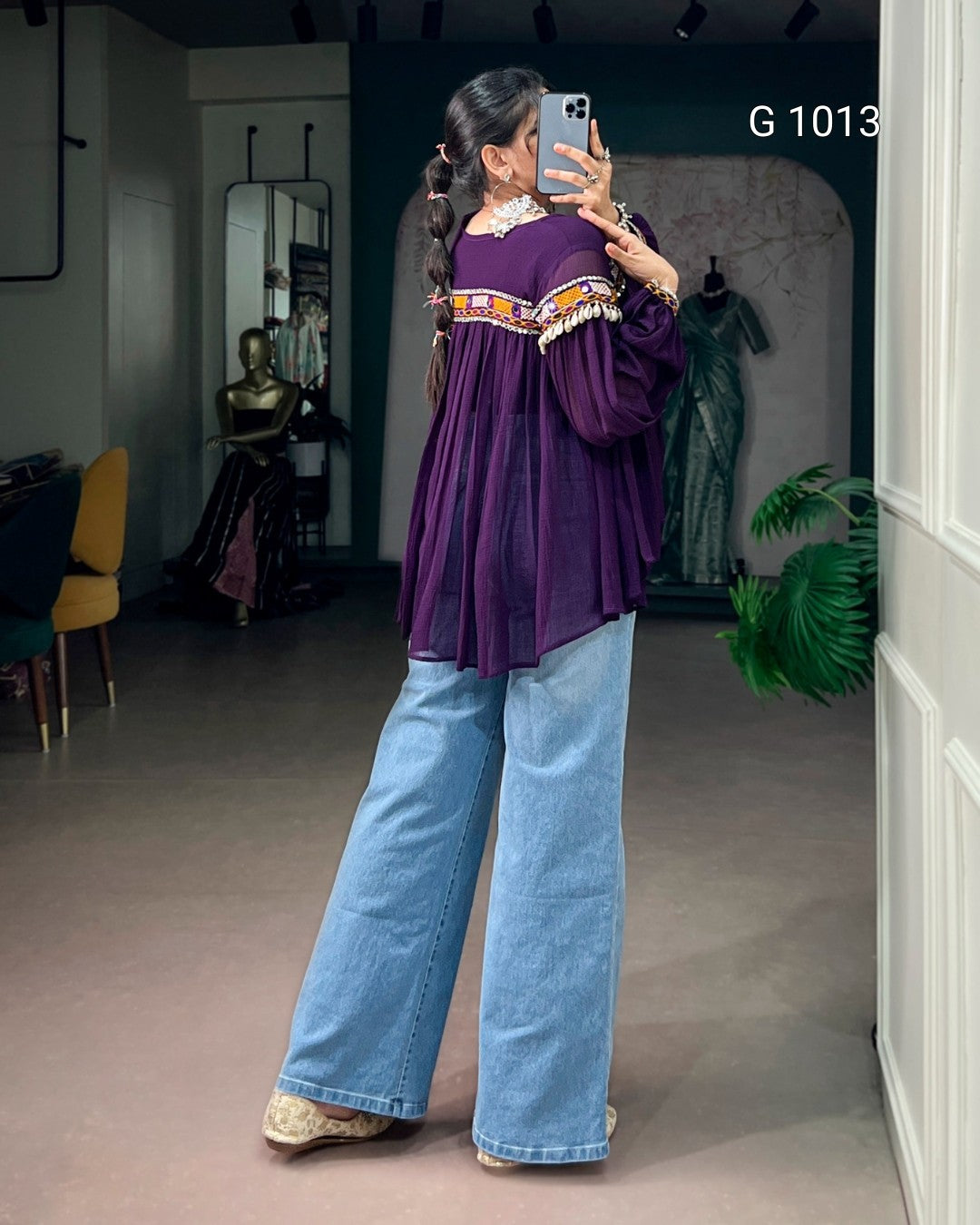 A woman takes a mirror selfie in a boutique, wearing a purple long-sleeve blouse with colorful embroidered details across the shoulders and light blue wide-leg jeans.