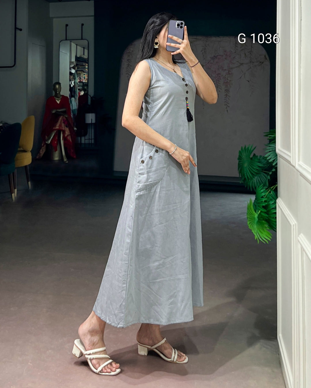 A woman takes a mirror selfie wearing a sleeveless, light gray midi dress with decorative buttons down the front and side pockets. She is wearing beige heeled sandals in a boutique setting.