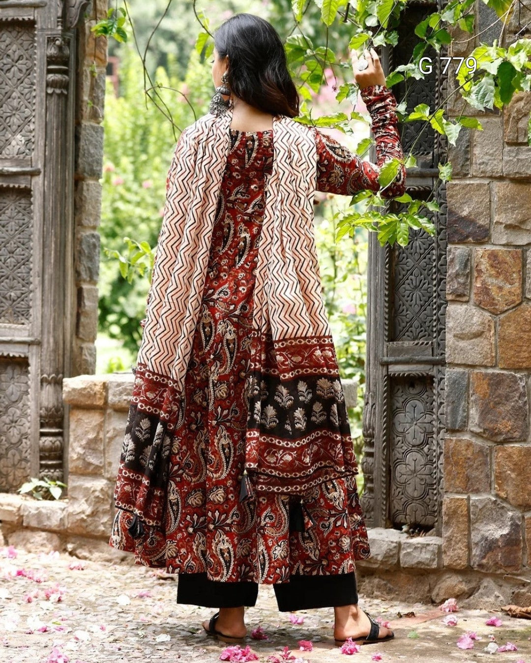 Woman in a patterned dress standing in a stone-walled garden with greenery.
