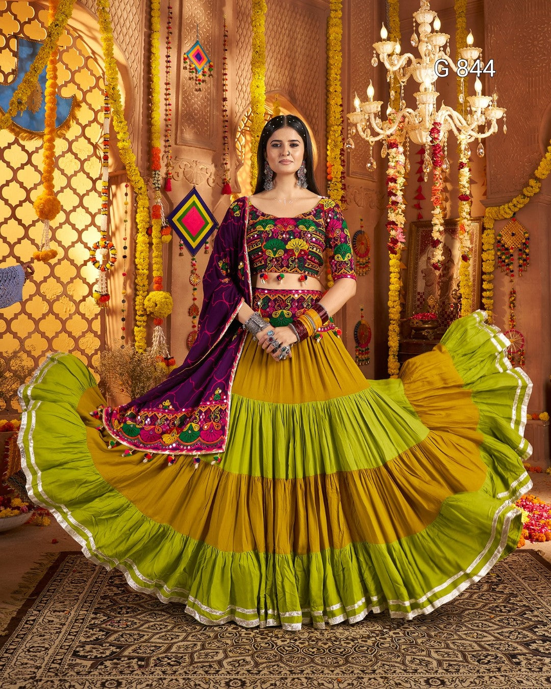 Woman in a traditional green and purple outfit with intricate patterns in a decorated indoor setting.