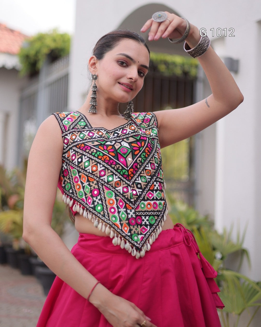 A woman wearing a sleeveless embroidered ethnic blouse with colorful geometric mirror-work patterns and tassel trim, paired with a bright pink skirt, posing outdoors in front of a white building.
