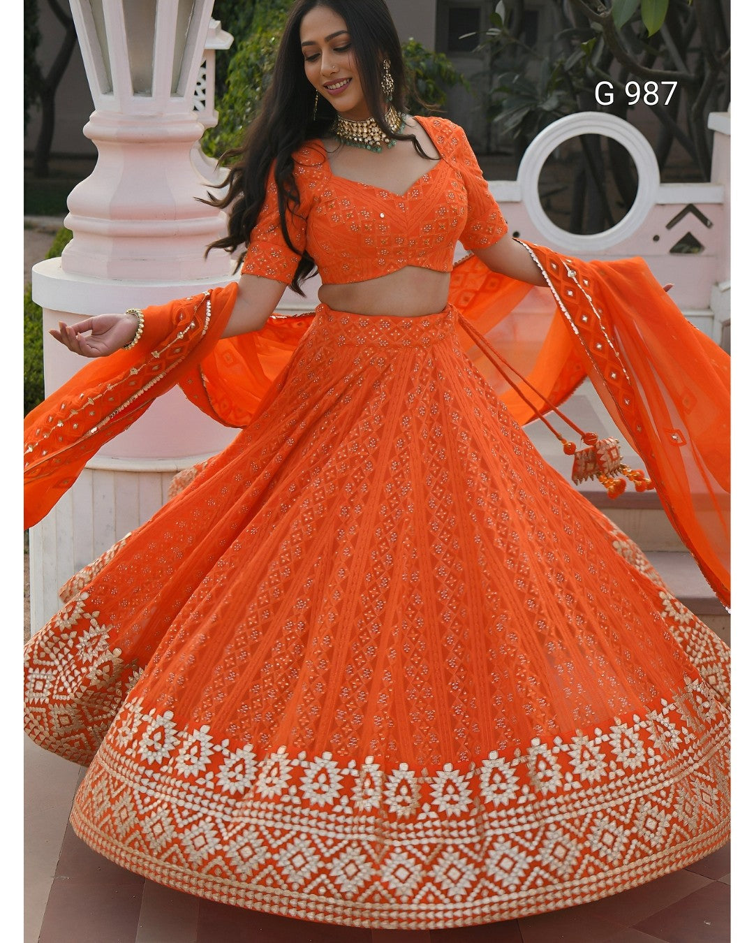 An orange embroidered Indian ethnic wear set consisting of a flared lehenga skirt, a matching crop top, and a dupatta, shown on a model. The set features intricate white and silver embroidery along the borders and bodice.