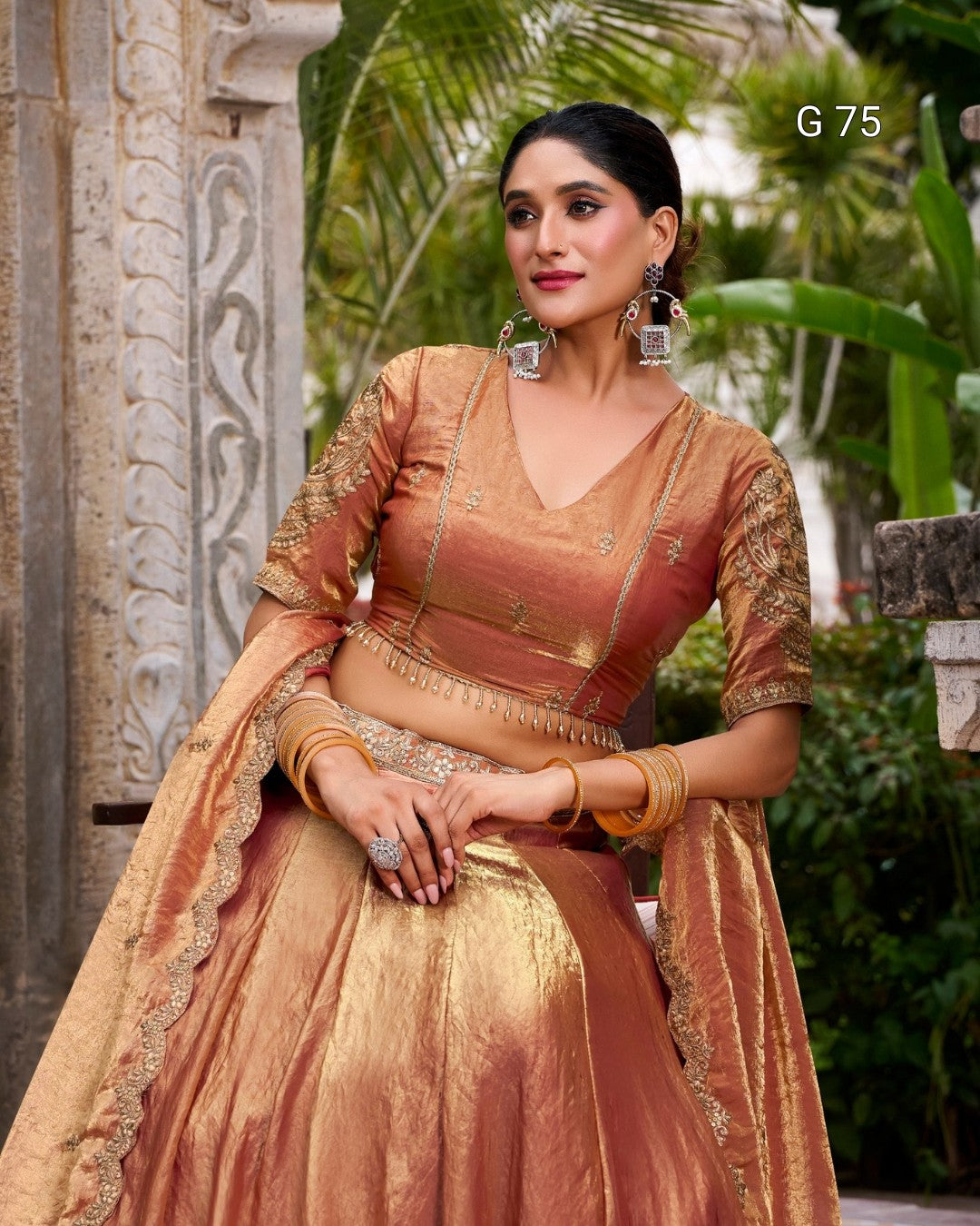 A woman wearing a traditional Indian salwar kameez outfit in a warm peach and gold color, with a matching dupatta, set against a background of stone pillars and green foliage. The text 'G 75' is visible in the top right corner.