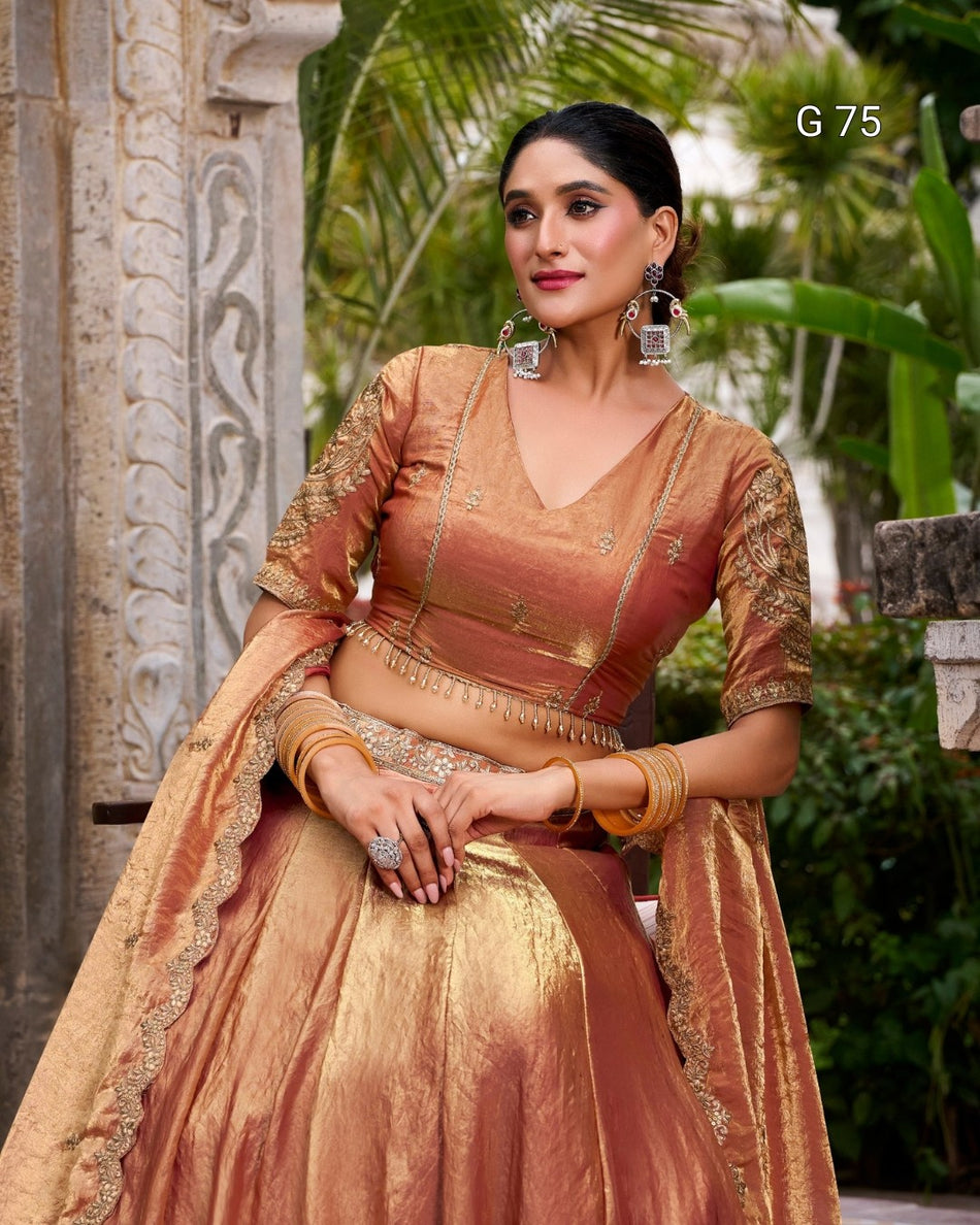 A woman wearing a traditional Indian salwar kameez outfit in a warm peach and gold color, with a matching dupatta, set against a background of stone pillars and green foliage. The text 'G 75' is visible in the top right corner.
