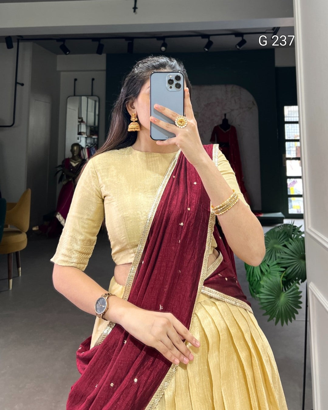 Woman taking a mirror selfie wearing a gold blouse and maroon saree in an indoor setting.
