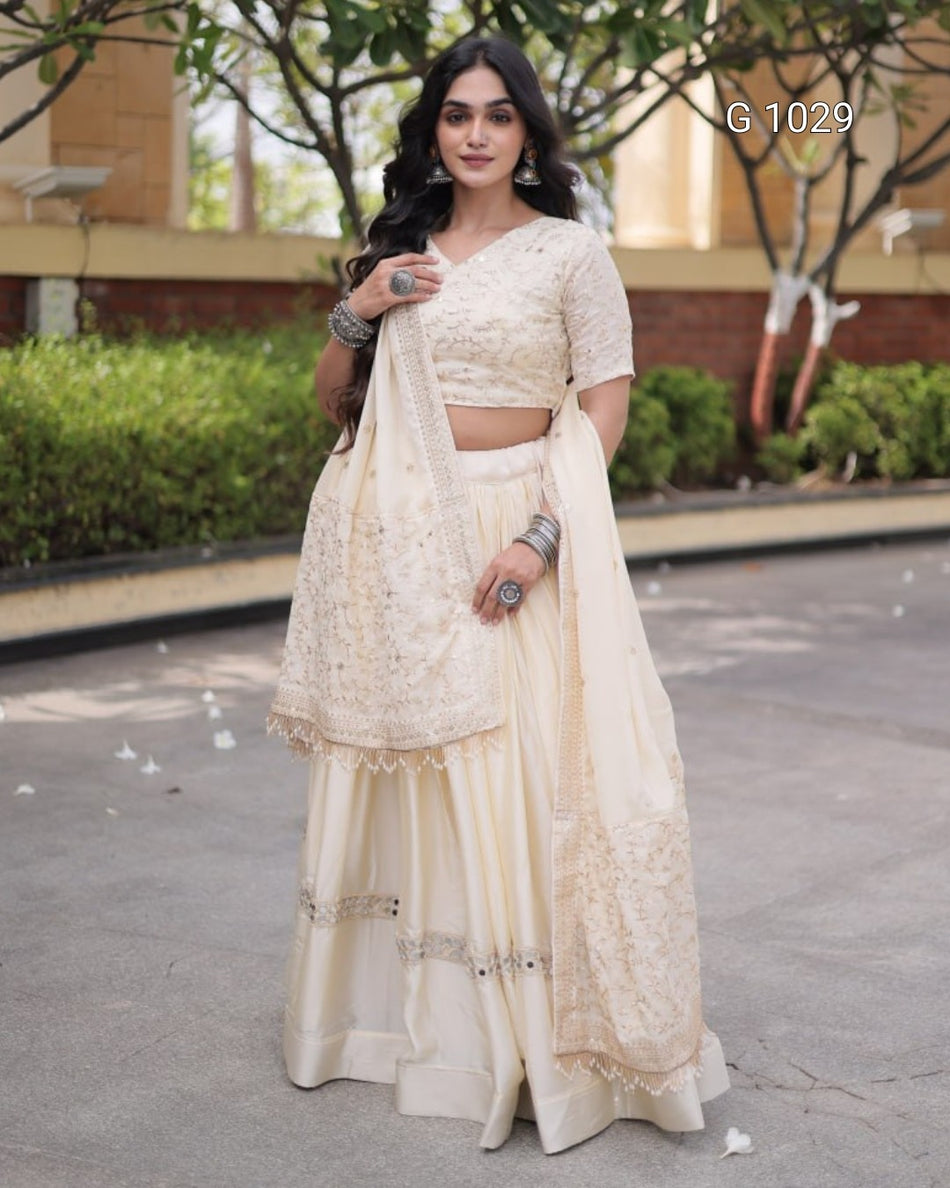 A woman models a cream-colored traditional Indian outfit consisting of a heavily embroidered crop blouse, a flared skirt, and a matching dupatta, set against an outdoor background with trees and a building.