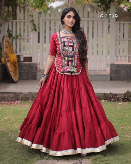 A woman wearing a traditional red Indian ethnic gown with a heavily embroidered bodice and a full tiered skirt, standing outdoors on a lawn in front of a white picket fence.