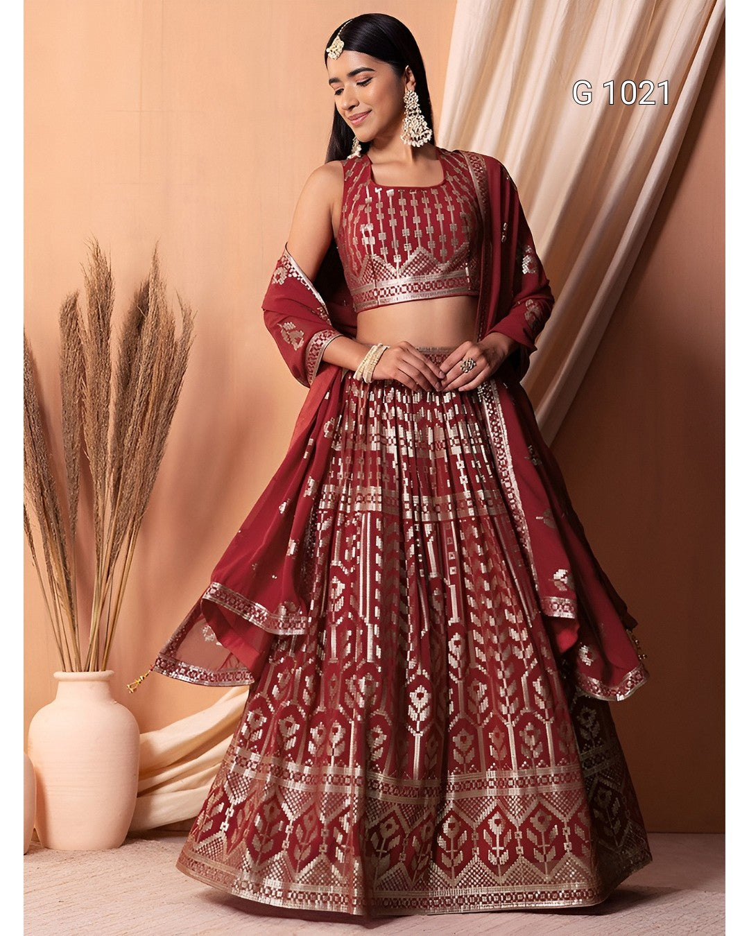 A woman models a traditional Indian lehenga choli set in deep maroon with intricate gold and silver metallic embroidery. The set includes a heavily embroidered crop top, a full flared skirt with geometric patterns, and a matching dupatta draped over her shoulders.