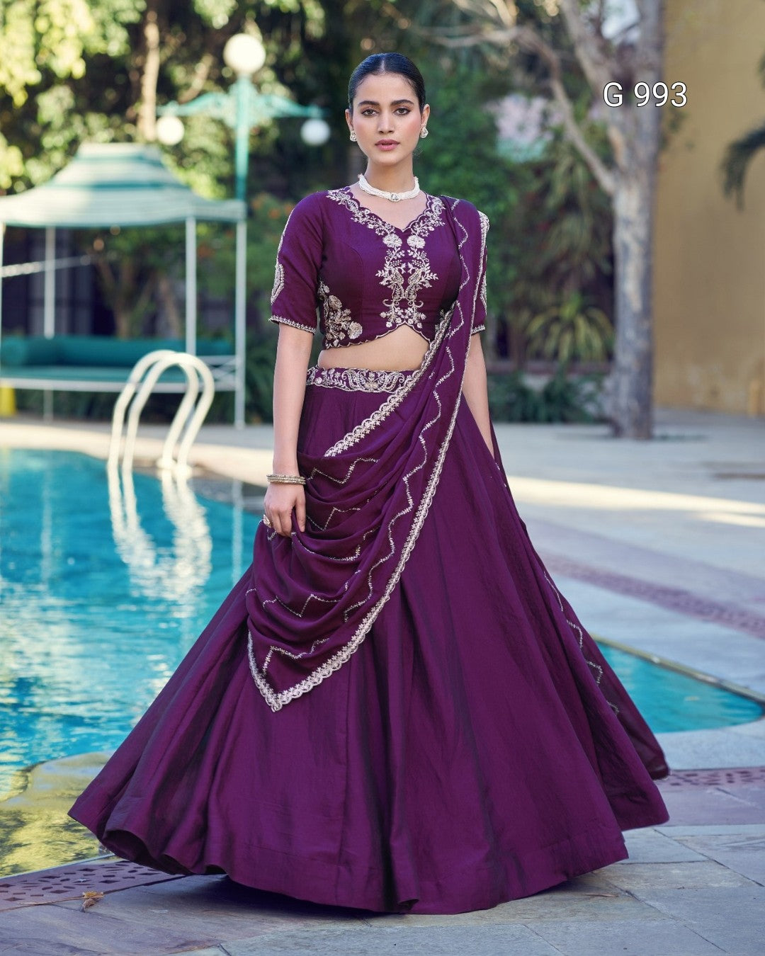 A woman stands outdoors beside a swimming pool wearing a deep purple traditional outfit with ornate gold embroidery. The outfit includes a flared skirt, fitted bodice, short sleeves, and a draped sash.