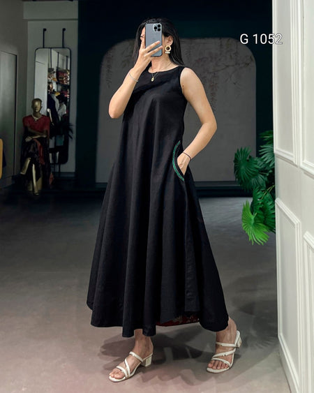 A woman takes a mirror selfie wearing a sleeveless black midi dress with a high neckline and side pockets, paired with white strappy heeled sandals. The background shows a boutique interior with a mannequin, mirror, and potted plant.
