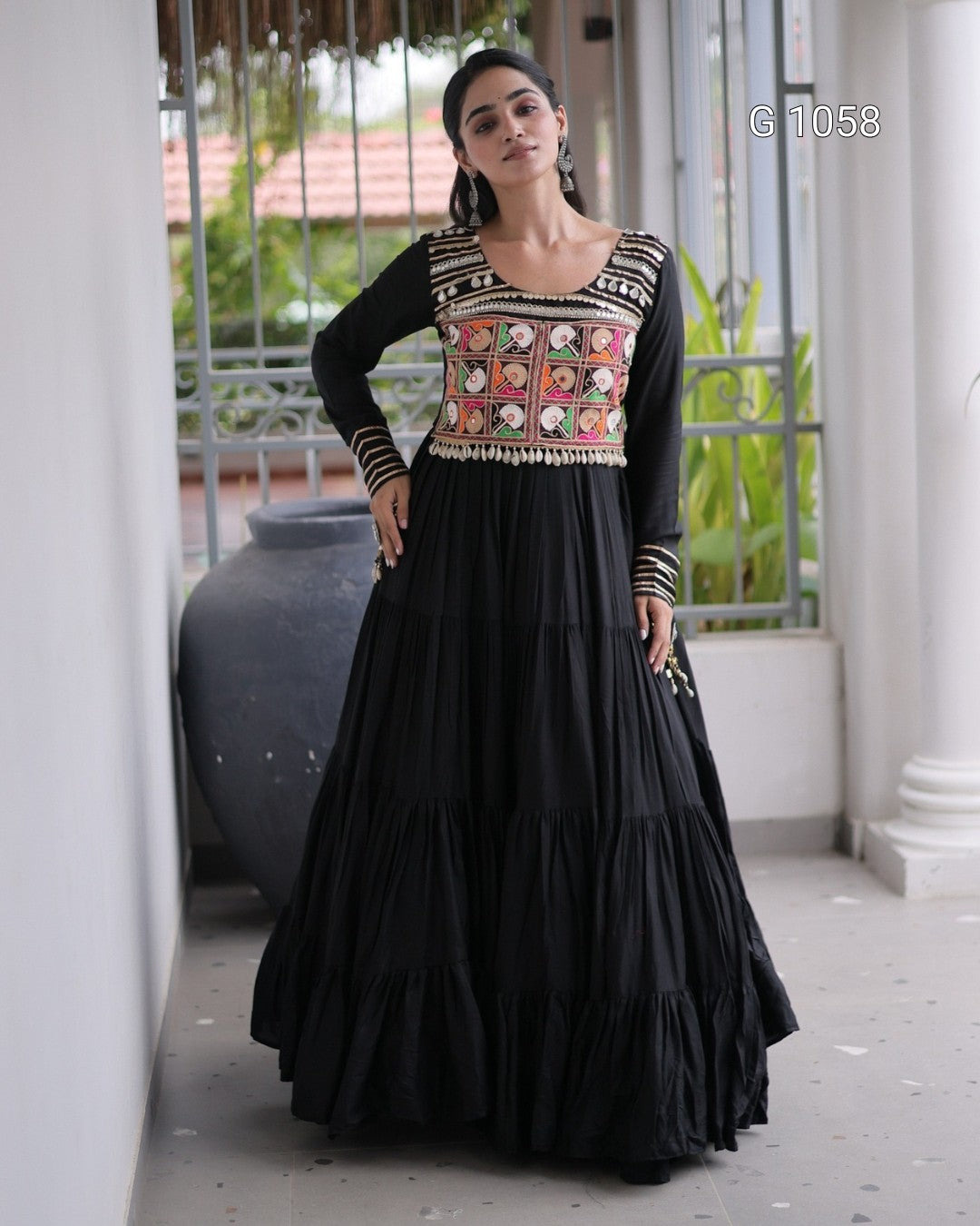 A woman models a black long-sleeve Anarkali gown with a heavily embroidered multicolor bodice featuring pink, orange, and gold patterns, set against a traditional corridor background.