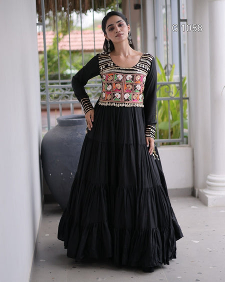 A woman models a black long-sleeve Anarkali gown with a heavily embroidered multicolor bodice featuring pink, orange, and gold patterns, set against a traditional corridor background.
