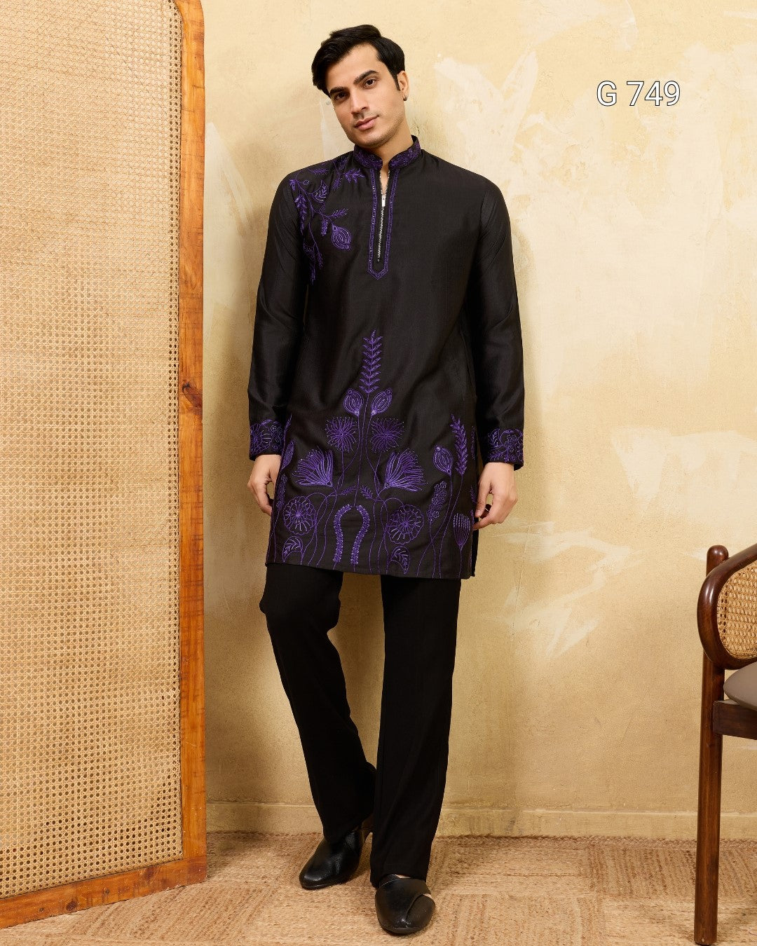 Man wearing a black kurta with purple embroidery against a beige wall.