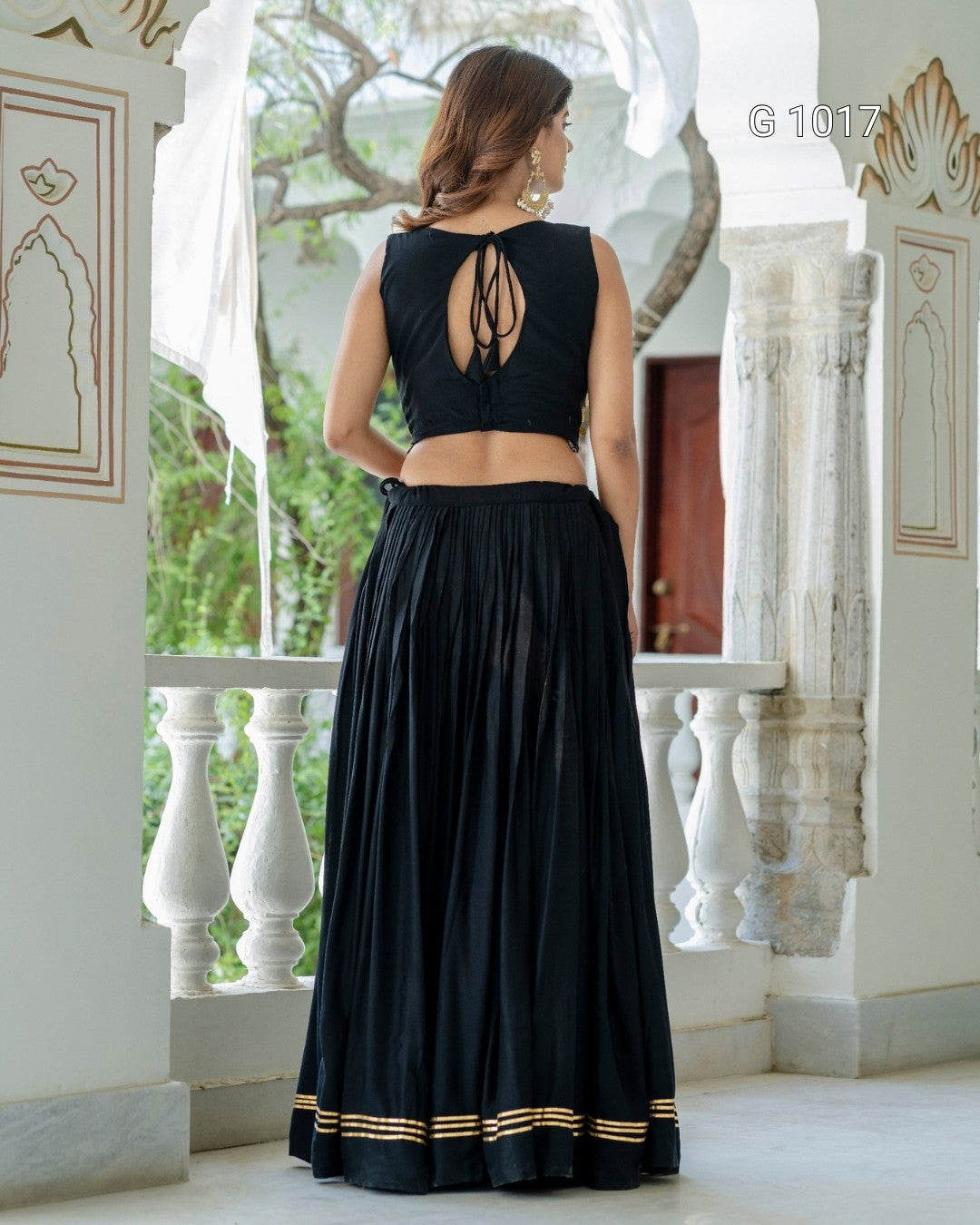 A woman stands with her back to the camera wearing a black sleeveless maxi dress with a tie-back neckline and a flared skirt. The dress has gold stripes near the hem and is photographed in an ornate indoor setting with white pillars and windows.
