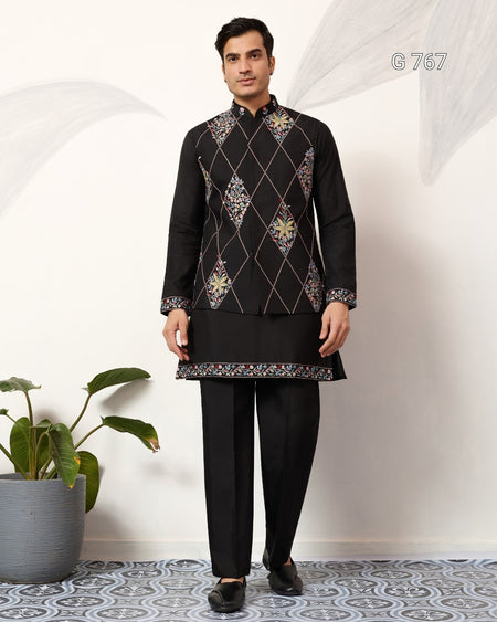 Man wearing a traditional black embroidered outfit standing indoors with a plant in the background.