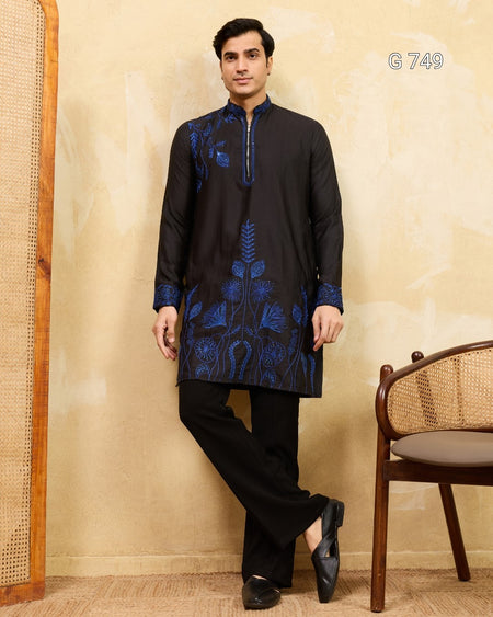 Man wearing a black kurta with blue embroidery standing against a beige wall.