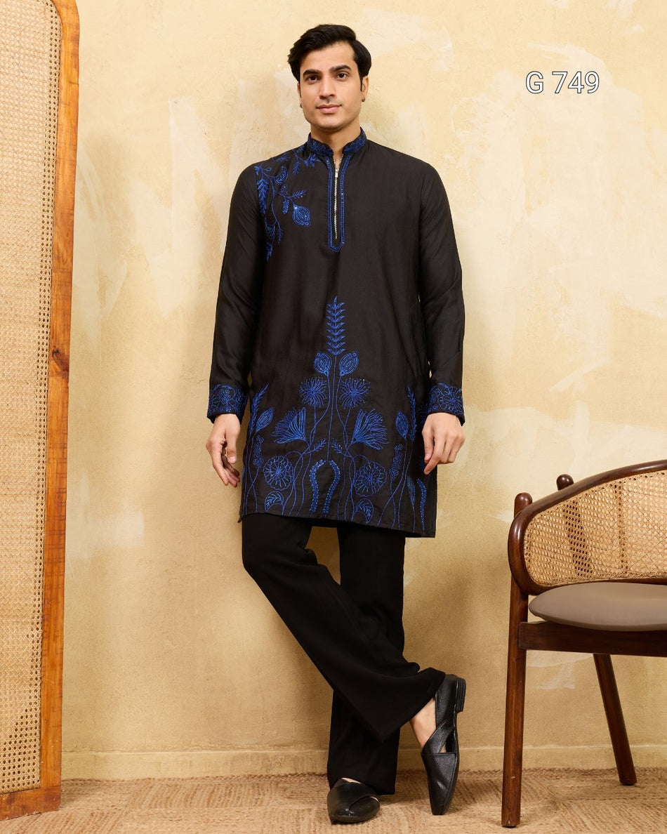 Man wearing a black kurta with blue embroidery standing against a beige wall.