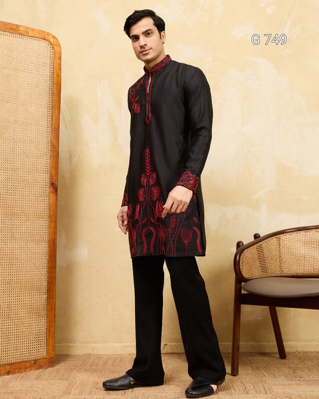Man wearing a black kurta with red embroidery in a room with a wooden chair and screen.