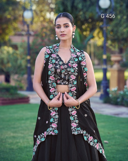 A woman stands outdoors in a garden wearing a black traditional outfit with colorful floral embroidery in pink, green, and blue. The outfit includes a sleeveless vest, matching embroidered crop top, and flared skirt.