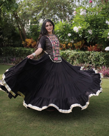A woman outdoors in a garden wearing a black traditional dress with a heavily embroidered multicolor bodice and a flared skirt with a cream border, captured mid-spin.