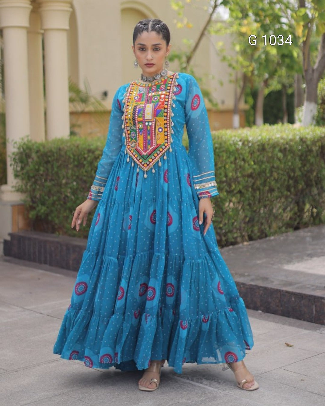 A woman modeling a bright blue traditional long dress with colorful embroidered details on the chest and scattered circular motifs on the skirt, standing outdoors in front of a building and hedges.