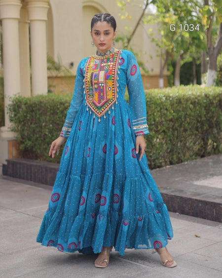 A woman modeling a bright blue traditional long dress with colorful embroidered details on the chest and scattered circular motifs on the skirt, standing outdoors in front of a building and hedges.