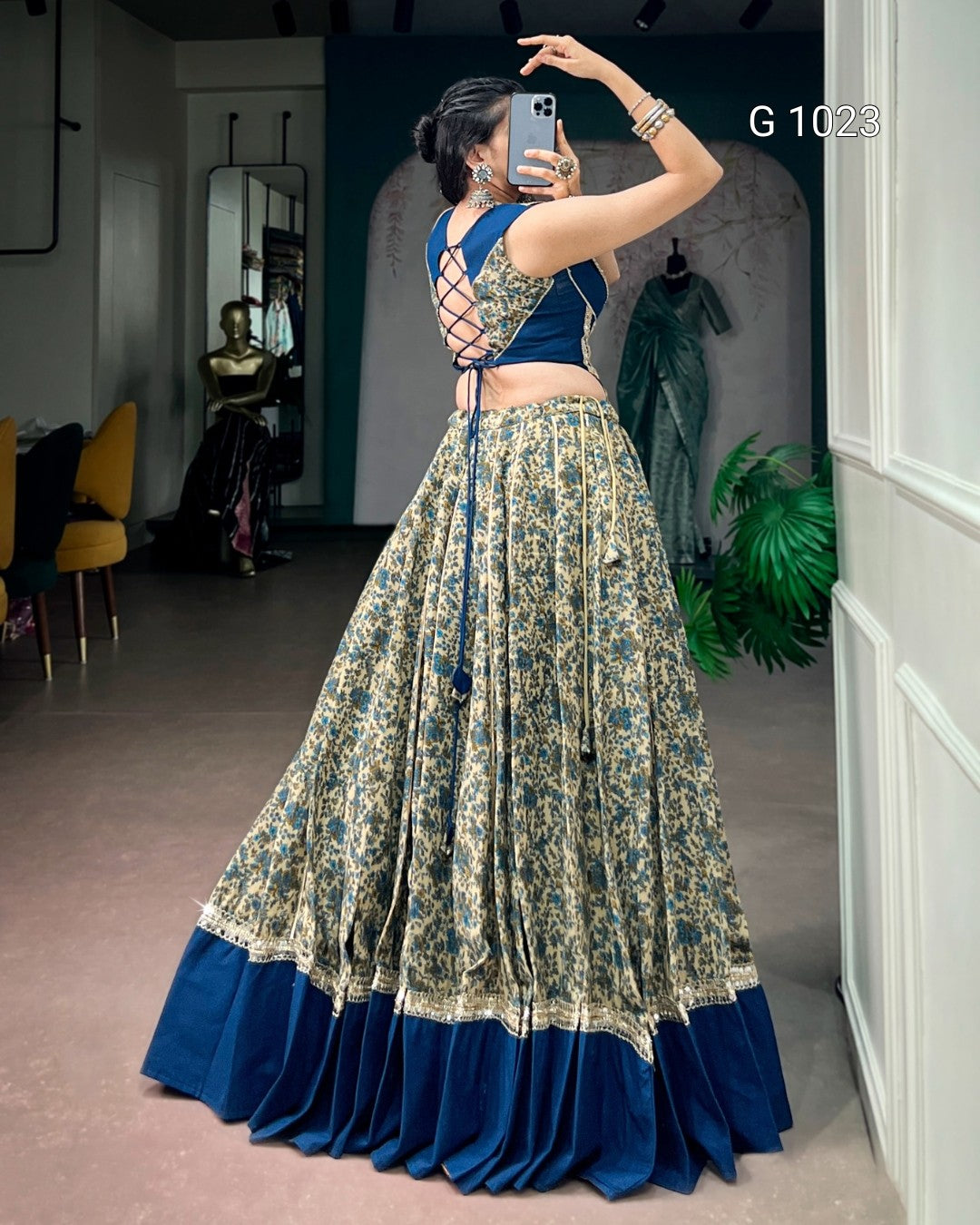 A woman takes a mirror selfie in a boutique wearing a traditional Indian lehenga choli. The outfit features a navy blue sleeveless choli with a criss-cross back design and a matching pleated skirt in beige and blue floral print with a solid navy blue border.