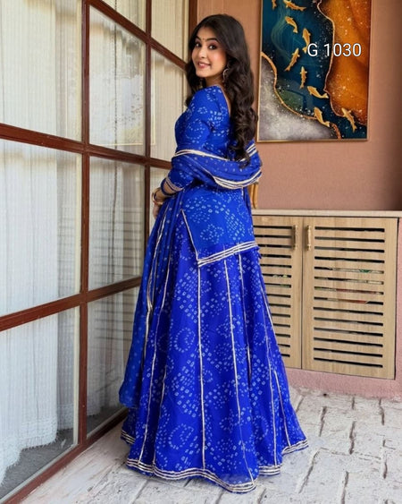 A woman modeling a royal blue traditional Indian dress with white bandhani tie-dye patterns and silver borders, standing in a decorated indoor setting with wooden window frames and a slatted cabinet.