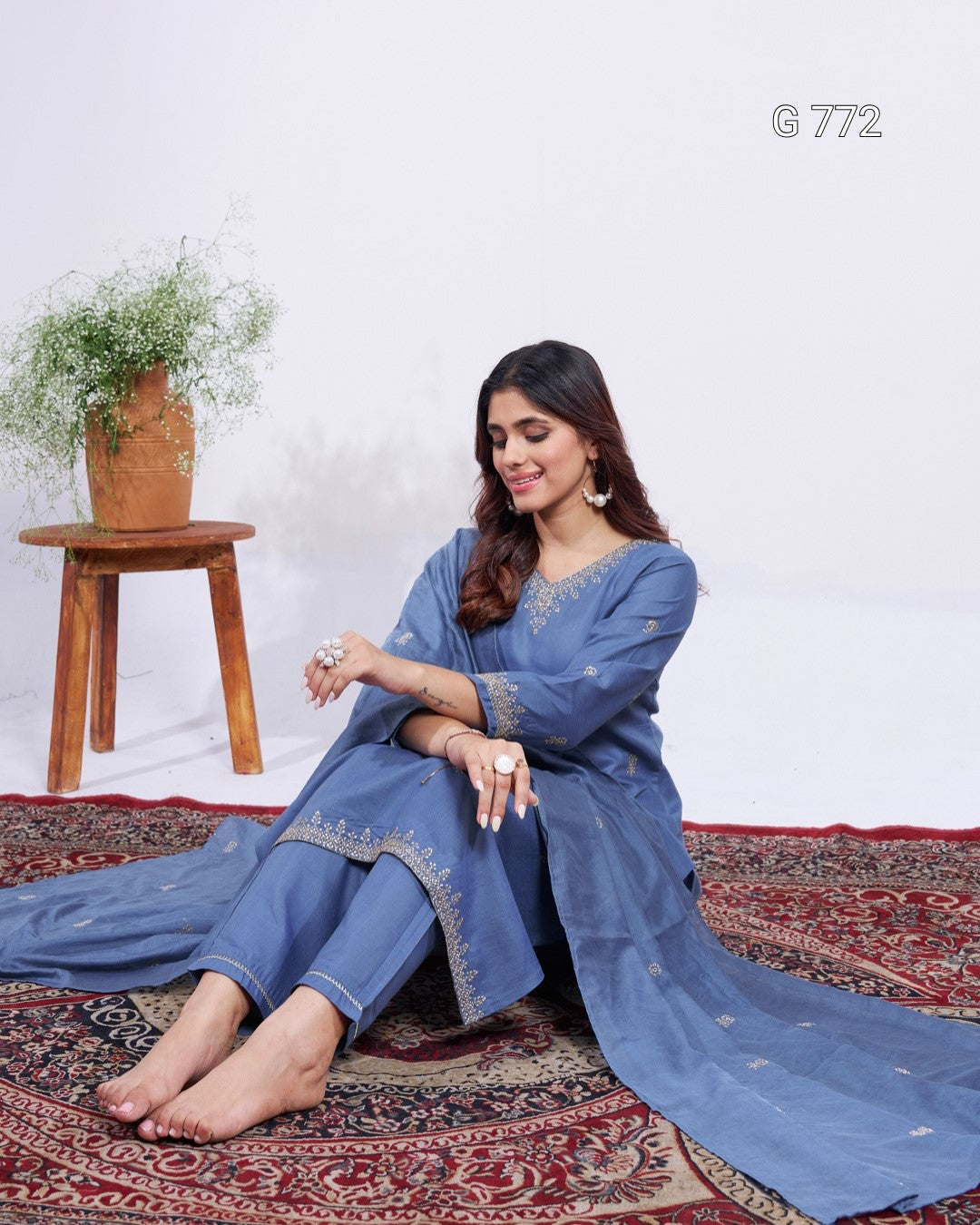 Woman in a blue traditional outfit sitting on a patterned rug with a wooden stool and plant in the background.
