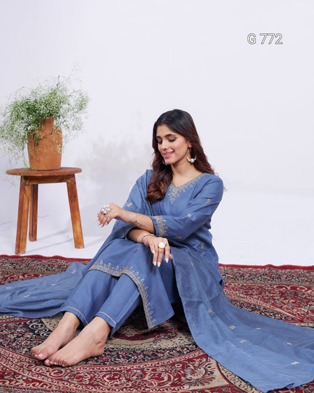 Woman in a blue traditional outfit sitting on a patterned rug with a wooden stool and plant in the background.