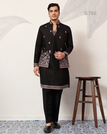 Man wearing a black traditional outfit with intricate patterns standing next to a wooden stool on a white background.